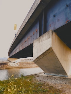 A large concrete and steel bridge spans over a body of water, with grass and plants growing along the banks. The underside of the bridge reveals structural details, and a sign with arrows is visible on the side. In the distance, there are some industrial buildings.
