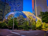 A modern urban landscape featuring two large geodesic dome structures surrounded by lush greenery. Tall office buildings with reflective glass facades rise in the background, and a streetlight is situated in the foreground. The sky is clear and blue, casting a vibrant light over the scene.
