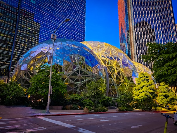 A modern urban landscape featuring two large geodesic dome structures surrounded by lush greenery. Tall office buildings with reflective glass facades rise in the background, and a streetlight is situated in the foreground. The sky is clear and blue, casting a vibrant light over the scene.