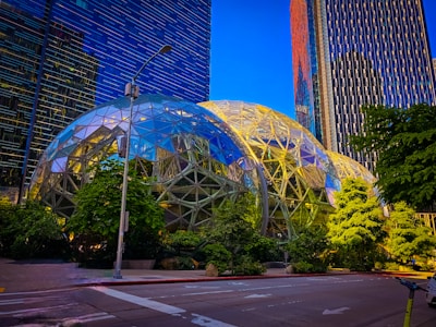 A modern urban landscape featuring two large geodesic dome structures surrounded by lush greenery. Tall office buildings with reflective glass facades rise in the background, and a streetlight is situated in the foreground. The sky is clear and blue, casting a vibrant light over the scene.