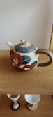 A ceramic teapot with a single spout and a sturdy handle. It features floral patterns in red and blue, surrounded by beige tones. The teapot is placed on a wooden surface, with two ceramic cups visible beneath it.