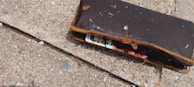 A worn-out brown wallet lying on a textured concrete surface. The wallet is partially open, revealing colorful cards inside, and is covered with dirt and small debris.