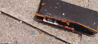 A worn-out brown wallet lying on a textured concrete surface. The wallet is partially open, revealing colorful cards inside, and is covered with dirt and small debris.