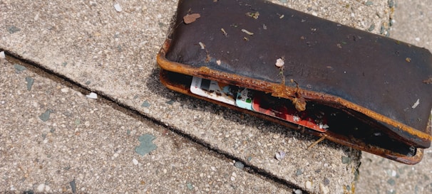 A worn-out brown wallet lying on a textured concrete surface. The wallet is partially open, revealing colorful cards inside, and is covered with dirt and small debris.