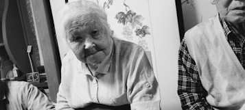An elderly woman with short white hair, wearing a striped shirt, is seated in a room with traditional decor. Next to her is an older man partially visible, wearing a checkered shirt and a vest. The background includes floral artwork and various objects on a shelf.