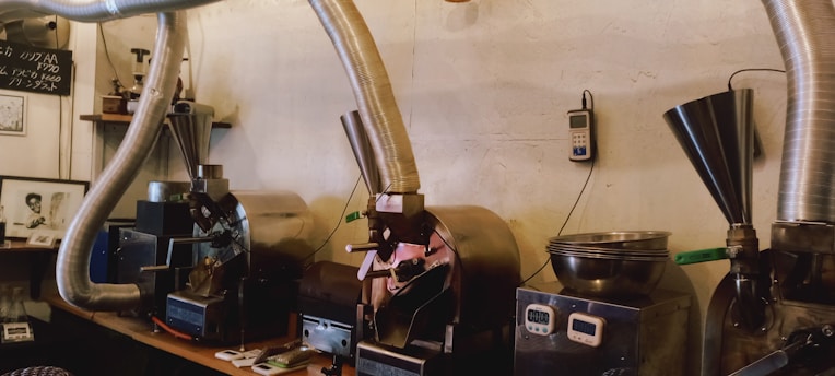 A row of industrial coffee roasting machines with metal ducting pipes attached to them. Various tools and small equipment are placed on the counter beside the machines. The setting appears to be in a workshop or small factory with a rustic, industrial decor.