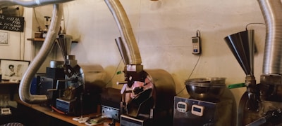 A row of industrial coffee roasting machines with metal ducting pipes attached to them. Various tools and small equipment are placed on the counter beside the machines. The setting appears to be in a workshop or small factory with a rustic, industrial decor.