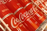 Rows of red Coca-Cola cans are displayed on a shelf, featuring the iconic white logo and the phrases 'Delicious & Refreshing' and 'Original'. The cans are slightly tilted, creating a visually appealing pattern.