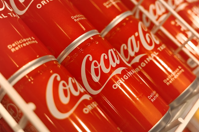 Rows of red Coca-Cola cans are displayed on a shelf, featuring the iconic white logo and the phrases 'Delicious & Refreshing' and 'Original'. The cans are slightly tilted, creating a visually appealing pattern.