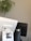 A close-up of a point-of-sale terminal with a visible logo sticker displaying contact details. A plant with long leaves is blurred in the background, and another device is also partially visible.