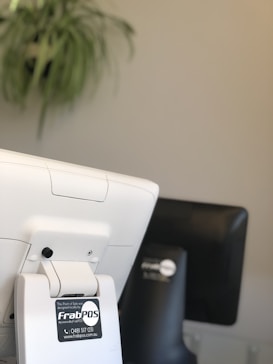A close-up of a point-of-sale terminal with a visible logo sticker displaying contact details. A plant with long leaves is blurred in the background, and another device is also partially visible.
