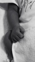 A close-up of a baby's hand resting on a soft, textured blanket. The baby is wearing a light-colored garment with some writing or design on it. The image is in grayscale, emphasizing the gentle and tender nature of a newborn's hand.