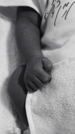 A close-up of a baby's hand resting on a soft, textured blanket. The baby is wearing a light-colored garment with some writing or design on it. The image is in grayscale, emphasizing the gentle and tender nature of a newborn's hand.