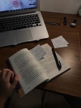 A workspace featuring a wooden desk with an open book written in Chinese, a pen resting on the pages, and several sticky notes with handwritten notes. A laptop is positioned in the upper left corner, displaying a video call. The desk also holds a watch, wristband, and a ring.