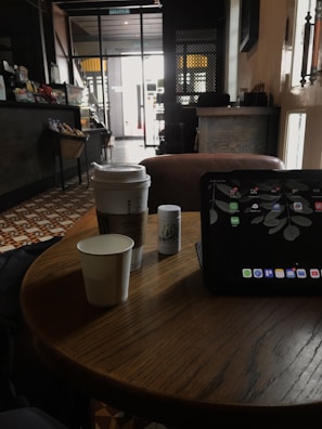 A cozy café setting featuring a small wooden table with a few items on top, including a cup with a lid, a paper cup, a small cylindrical container, and a tablet displaying a leaf-themed wallpaper. The background consists of a patterned tile floor, a counter with snacks, and a glass door leading outside, allowing natural light to filter in.