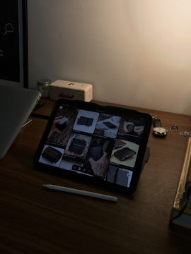 A dimly lit desk with an iPad displaying a Pinterest-like app. The iPad shows a variety of wallet images on its screen. Next to the iPad is a stylus, a closed laptop, a small speaker, and a wristwatch. The overall setting appears to be a workspace with minimal lighting.