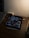 A dimly lit desk with an iPad displaying a Pinterest-like app. The iPad shows a variety of wallet images on its screen. Next to the iPad is a stylus, a closed laptop, a small speaker, and a wristwatch. The overall setting appears to be a workspace with minimal lighting.