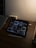 A dimly lit desk with an iPad displaying a Pinterest-like app. The iPad shows a variety of wallet images on its screen. Next to the iPad is a stylus, a closed laptop, a small speaker, and a wristwatch. The overall setting appears to be a workspace with minimal lighting.