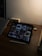 A dimly lit desk with an iPad displaying a Pinterest-like app. The iPad shows a variety of wallet images on its screen. Next to the iPad is a stylus, a closed laptop, a small speaker, and a wristwatch. The overall setting appears to be a workspace with minimal lighting.