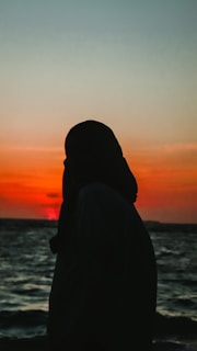 A silhouette of a person wearing a headscarf is set against a vibrant sunset sky with hues of orange and red. The ocean stretches out beneath the dramatic sky, with gentle waves visible.