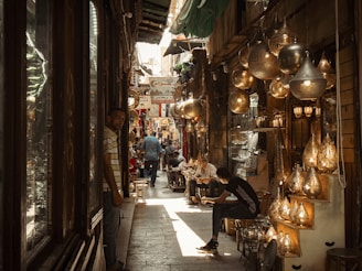 A bustling narrow marketplace with various hanging lanterns and lamps illuminating the scene. People are walking and sitting, creating an atmosphere of lively commerce and cultural exchange. The setting appears to be a traditional bazaar with a rich display of crafted metal goods.
