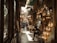 A bustling narrow marketplace with various hanging lanterns and lamps illuminating the scene. People are walking and sitting, creating an atmosphere of lively commerce and cultural exchange. The setting appears to be a traditional bazaar with a rich display of crafted metal goods.