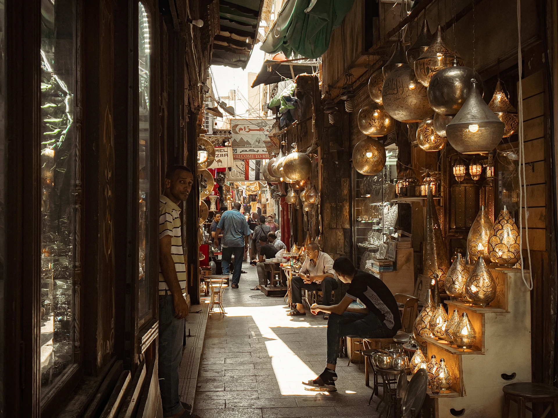 A bustling narrow marketplace with various hanging lanterns and lamps illuminating the scene. People are walking and sitting, creating an atmosphere of lively commerce and cultural exchange. The setting appears to be a traditional bazaar with a rich display of crafted metal goods.