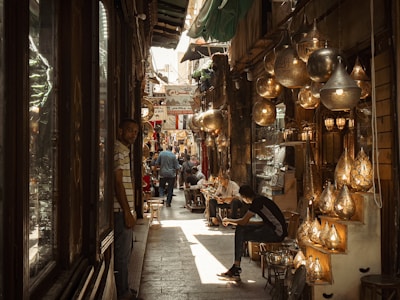 A bustling narrow marketplace with various hanging lanterns and lamps illuminating the scene. People are walking and sitting, creating an atmosphere of lively commerce and cultural exchange. The setting appears to be a traditional bazaar with a rich display of crafted metal goods.