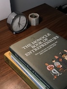 A wooden table features a small analog clock with a silver frame, a small concrete planter with a succulent, and a stack of three books titled 'The Monocle Book of Entrepreneurs', 'The Forecast', and 'The Monocle Guide to Better Living'. The top book has an illustration of people standing in a line.