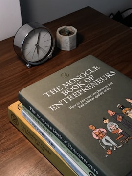 A wooden table features a small analog clock with a silver frame, a small concrete planter with a succulent, and a stack of three books titled 'The Monocle Book of Entrepreneurs', 'The Forecast', and 'The Monocle Guide to Better Living'. The top book has an illustration of people standing in a line.