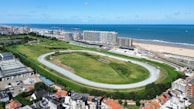 An aerial view depicts a large horse racing track surrounded by green fields and urban buildings. In the background, the ocean and a sandy beach stretch along the coast. Several cranes are present among the modern buildings, indicating ongoing construction. The area is vibrant with a mix of natural and urban elements, under a clear blue sky with scattered clouds.