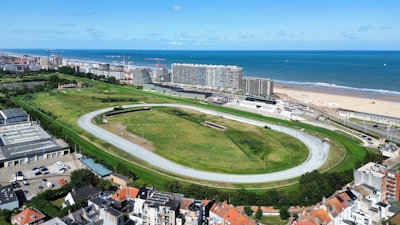 An aerial view depicts a large horse racing track surrounded by green fields and urban buildings. In the background, the ocean and a sandy beach stretch along the coast. Several cranes are present among the modern buildings, indicating ongoing construction. The area is vibrant with a mix of natural and urban elements, under a clear blue sky with scattered clouds.