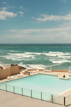 A calm seaside view with an inviting swimming pool in the foreground, surrounded by a glass barrier. The ocean beyond has gentle waves under a partly cloudy sky, creating a serene atmosphere.