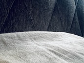 A close-up view of a textured gray fabric surface partially covered by a light, soft material. The background features a dark, patterned upholstery with diagonal stitching.