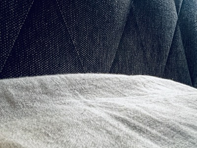 A close-up view of a textured gray fabric surface partially covered by a light, soft material. The background features a dark, patterned upholstery with diagonal stitching.