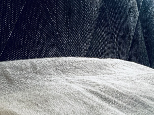 A close-up view of a textured gray fabric surface partially covered by a light, soft material. The background features a dark, patterned upholstery with diagonal stitching.
