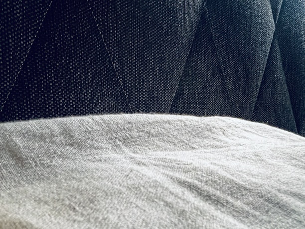 A close-up view of a textured gray fabric surface partially covered by a light, soft material. The background features a dark, patterned upholstery with diagonal stitching.