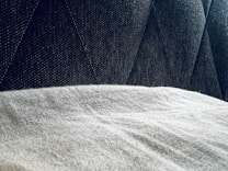 A close-up view of a textured gray fabric surface partially covered by a light, soft material. The background features a dark, patterned upholstery with diagonal stitching.