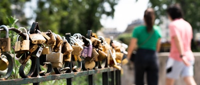 A collection of various padlocks attached to a metal fence, symbolizing love and commitment. In the background, two blurred figures, a man and a woman, are walking together, emphasizing a sense of togetherness.