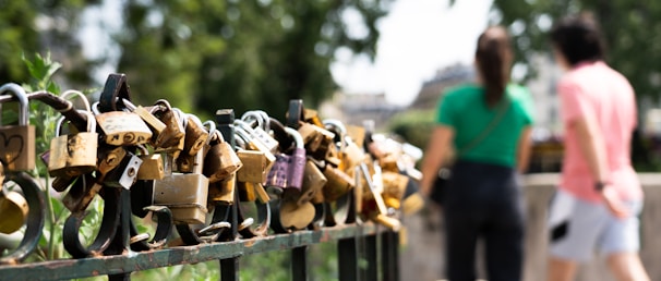 A collection of various padlocks attached to a metal fence, symbolizing love and commitment. In the background, two blurred figures, a man and a woman, are walking together, emphasizing a sense of togetherness.