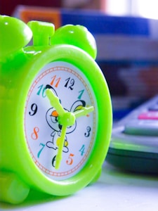 A small, vibrant green plastic clock with cartoon characters on its face. The clock features colorful numbers and yellow hands. It is placed next to a calculator on a white surface, with a blurred background showing blue and orange colors.