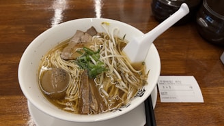 A bowl of ramen with noodles, slices of meat, bean sprouts, and chopped green onions, served in a light broth. A white spoon is placed on the rim of the bowl, and there's a soft-boiled egg inside the broth. A piece of paper with text is visible on the wooden table beside the bowl.