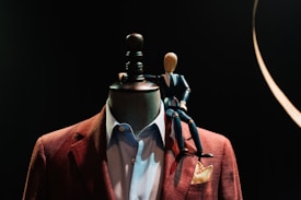 A mannequin adorned with a stylish brown blazer and shirt is topped with a wooden artist's mannequin sitting on its shoulder. The scene is set against a dark background, highlighting the contrast between the mannequin and its attire.
