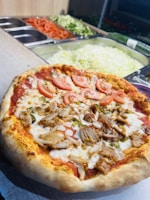A freshly cooked pizza with toppings of sliced tomatoes and shredded meat is positioned on a counter. Behind the pizza, there is a preparation station with containers holding sliced tomatoes, shredded lettuce, cucumbers, and other vegetables.