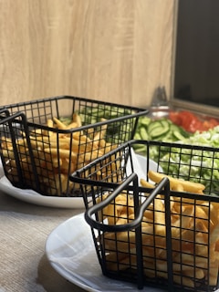 Two black metal baskets filled with golden, crispy French fries are placed on white plates. In the background, there is a wooden surface, and a colorful salad featuring sliced cucumbers, tomatoes, and greens.