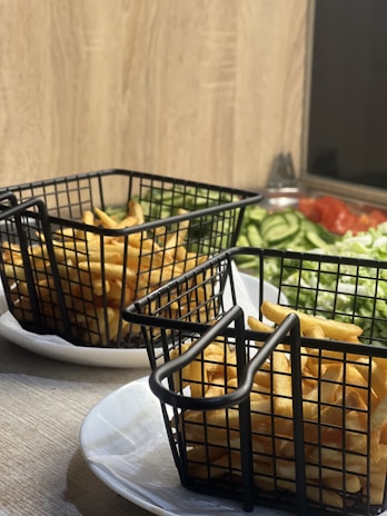 Two black metal baskets filled with golden, crispy French fries are placed on white plates. In the background, there is a wooden surface, and a colorful salad featuring sliced cucumbers, tomatoes, and greens.