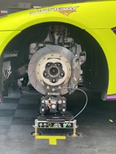 A detailed view of a high-performance vehicle's wheel section without the tire, showcasing the complex brake disc and caliper system mounted on an advanced setup tool. The car's body is partially visible, painted in bright green with racing decals. The focus is on the meticulously detailed brake components and the equipment used for servicing or testing.