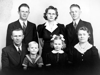 A black and white vintage photograph of a family with seven members, including four adults and three children, all dressed in formal attire. The adults are wearing suits and dresses typical of mid-20th century fashion, while the children are in neatly styled outfits. The group is posed in a traditional manner, likely for a formal family portrait.