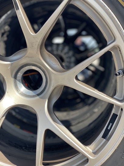 A close-up view of a metallic car wheel rim with a five-spoke design. The surface shows some wear, and the background reveals a blurred image with more wheels and indistinct objects.
