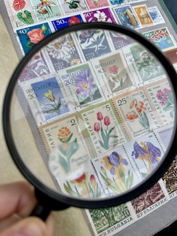 A collection of colorful postage stamps adorned with floral illustrations is magnified by a handheld magnifying glass. Each stamp features different types of flowers with a variety of vibrant colors and intricate details. The hand holding the magnifying glass is visible at the edge of the image.